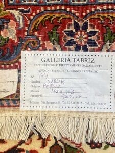 Un primo piano di un'etichetta su un tappeto sarouk persiano che riporta in dettaglio le informazioni sulla vendita e sul restauro, posizionata sullo sfondo fantasia del tappeto, funge da bozza automatica per mostrare i dettagli intricati.