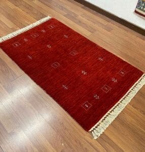 Galleria Tabriz - Tappeti in vendita. Su un pavimento in legno è adagiato un tappeto rosso rettangolare con nappe bianche su due estremità, decorato con piccoli motivi geometrici e vegetali di Bozza automatica.