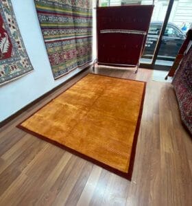 Galleria Tabriz - Tappeti in vendita. In una stanza con pavimento in legno è esposto un tappeto arancione bordato di marrone, circondato da vari tappeti a fantasia appesi alle pareti, creando un gioco di colori e consistenze.