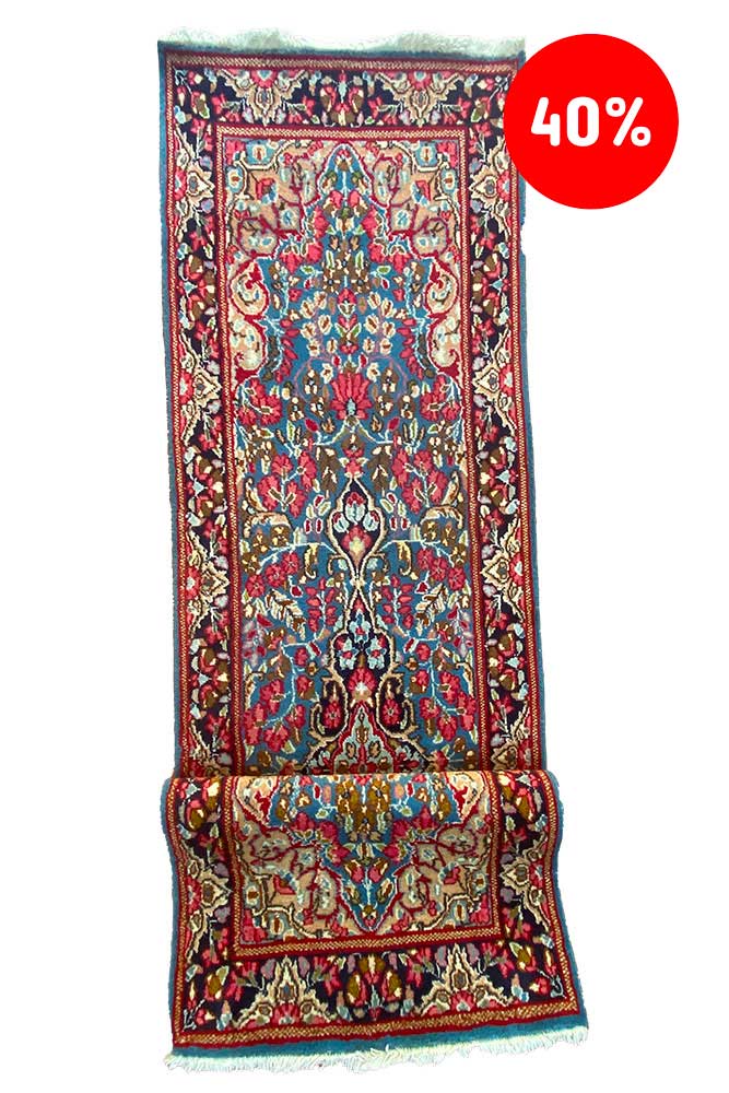 Galleria Tabriz - Tappeti in vendita. Questo intricato tappeto Kirman รจ esposto verticalmente, esibendo elaborati motivi floreali in un accattivante schema cromatico rosso e blu. Un'etichetta circolare rossa nell'angolo mostra con orgoglio uno sconto del "40%", rendendo questo pezzo un'aggiunta lussuosa ma conveniente a qualsiasi spazio.