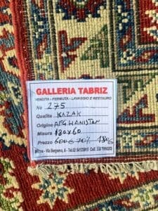 Galleria Tabriz - Tappeti in vendita. Primo piano di un tappeto con etichetta che riporta: Gallery Tabriz, tappeto kazako dall'Afghanistan, misura 180x60 cm, prezzo 500 euro. L'etichetta include un numero di telefono e un indirizzo.