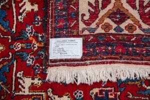 Galleria Tabriz - Tappeti in vendita. Primo piano dell'angolo di un tappeto persiano con etichetta Galleria Tabriz e frangia bianca su sfondo rosso, che ricorda i classici disegni Tappeto Heriz.