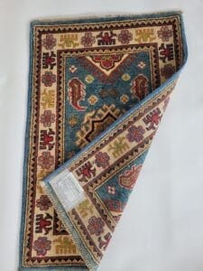 Galleria Tabriz - Tappeti in vendita. Un tappeto colorato e decorato giace parzialmente piegato su uno sfondo chiaro. Presenta disegni geometrici rossi, blu e beige, che rivelano un'etichetta sul retro.